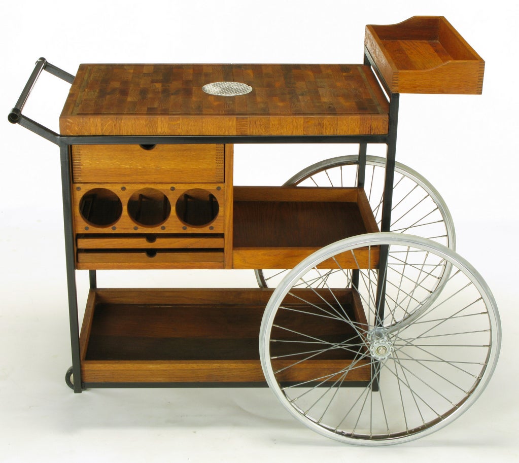 White oak and mahogany three wheel serving cart by Arthur Umanoff. Black lacquered iron frame, large bicycle style wheels and a single pivoting wheel. Butcher block top with center polished aluminum textured disc. Three wine bottle holder and as