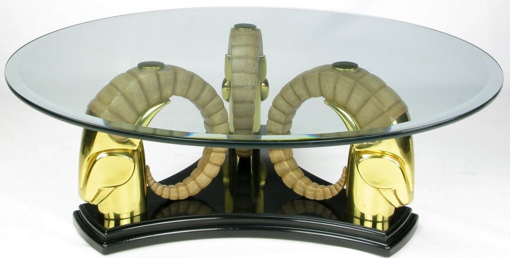 An elegant coffee table, with a nod to Chervet, featuring a trio of stylized ram heads in solid brass with individual resin horns anchored by a black lacquered wood base. The round, ogee edge glass top is 1/2
