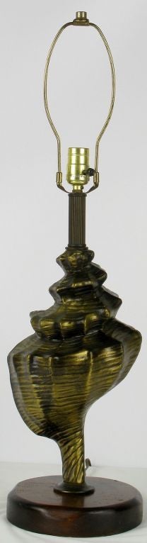 An unusual form for a table lamp, this cast metal conch shell is finished in a patinated gold with a carved wood base. A wonderful and unique table lamp that is possibly a Chapman design. Sold sans shade.