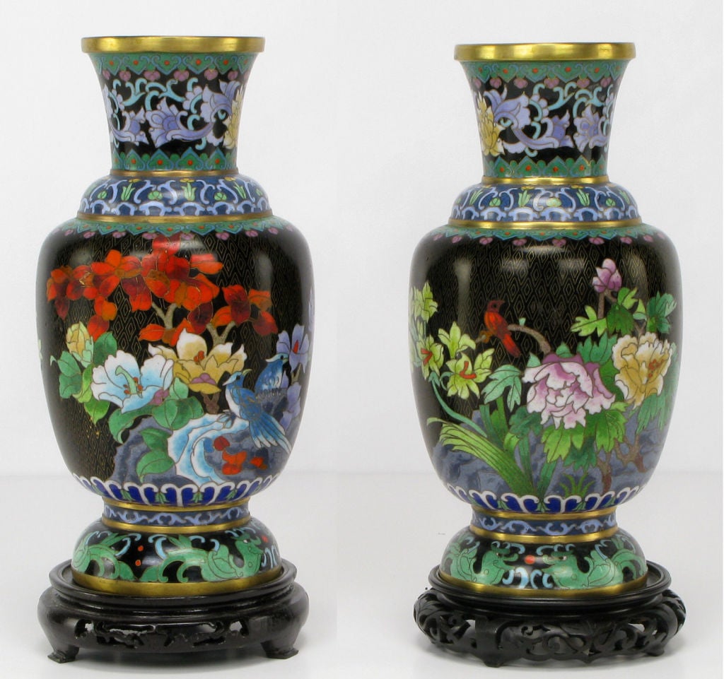 Pair of Chinese floral-decorated and mirror-image black cloisonne vases. Colorful flora and fauna depicted in green, red, blue, gold, purple and turquoise. Carved and black lacquered wood bases.