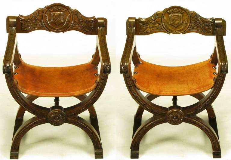 Pair of Spanish revival curule chairs in carved oak and umber leather with large brass nail head upholstery tacks. Hand carved backs feature square rosettes and center shields. X-shape sled bases feature center rosettes and carved stretchers.