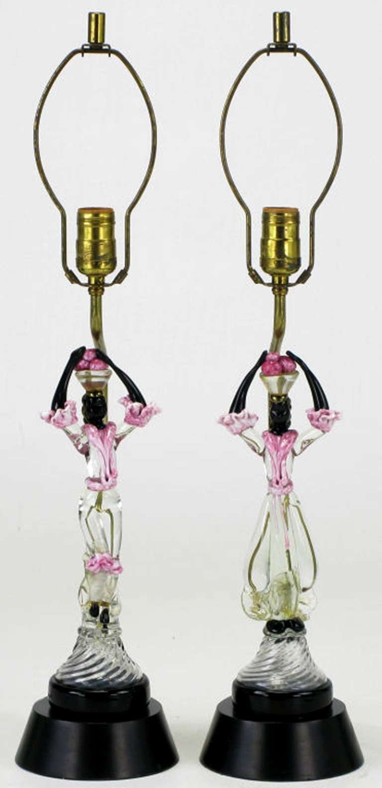 Pair of table lamps with fused Murano glass sculptures depicting African women, carrying baskets of fruit in colorful attire. Mounted on black lacquered bases with brass stems and sockets. Sold sans shades.