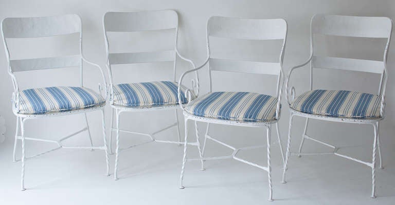 A sturdy set of four iron garden armchairs from France circa 1930. The chairs have open ironwork seats all in good condition and custom made cushions in a blue striped fabric. They are wide and comfortable enough for anyone to sit in.