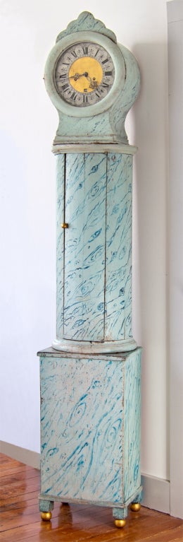 A Swedish tall clock in a rare pillar design with and original faux painting resembling marble. The face is an unusual combination of metal exterior with a gilded center echoing the gilt paint on the feet. The clock is in working order with the