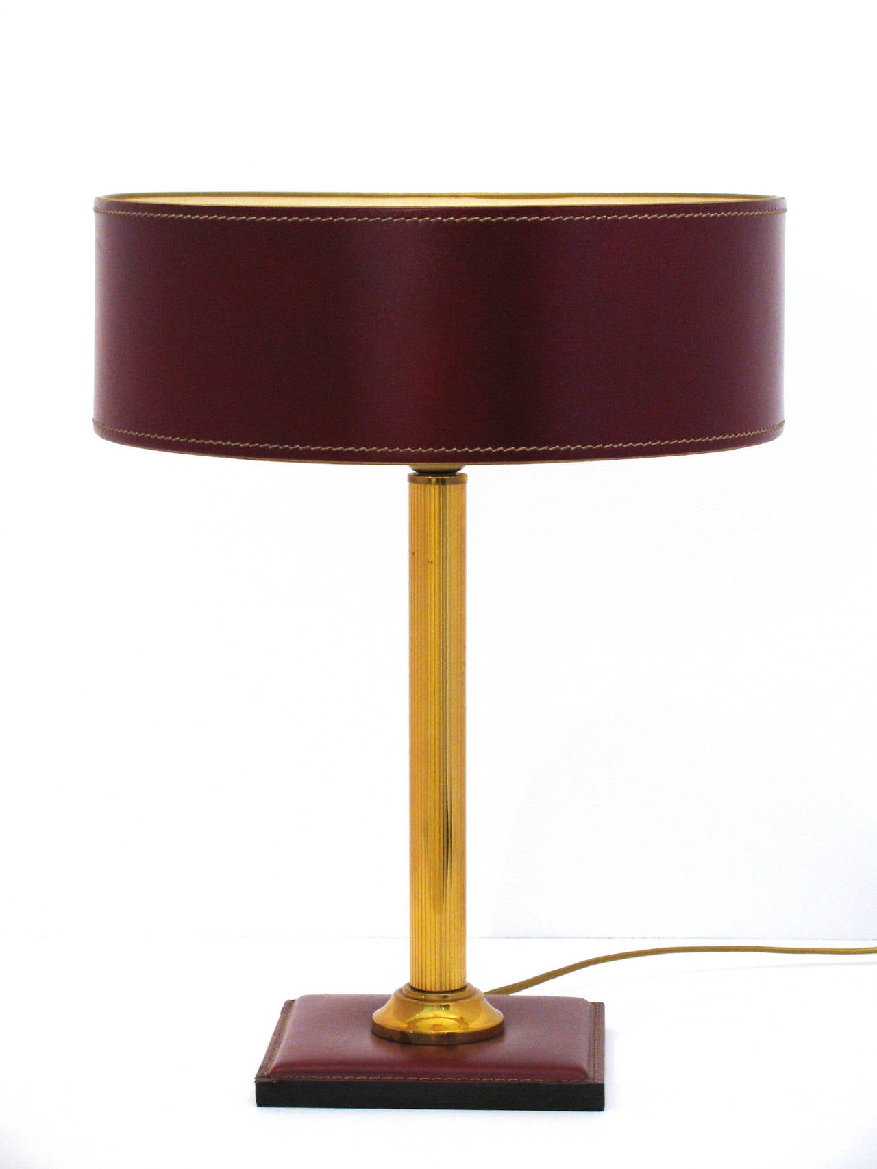 A classical bordeaux color leather-clad table lamp by Jacques Adnet. This stunning lamp features a hand-stitched shade with ivory paper lining, polished brass reeded column stem, and square base. On/off switch located on electrical cord. Please