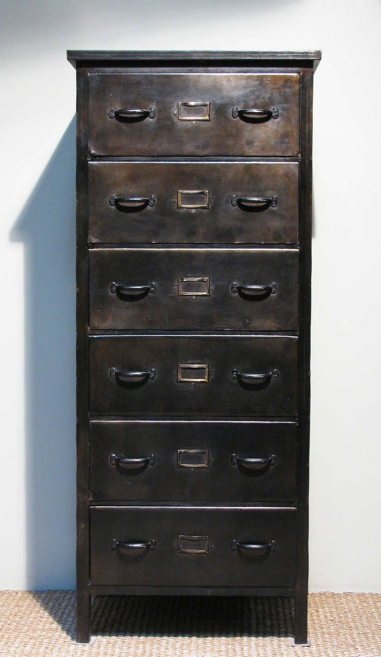 A tall rectangular metal chest with six full drawers; each fitted with double handles centered by slide-in label holder, raised on slender squared legs. Finish is a deep umber shade.