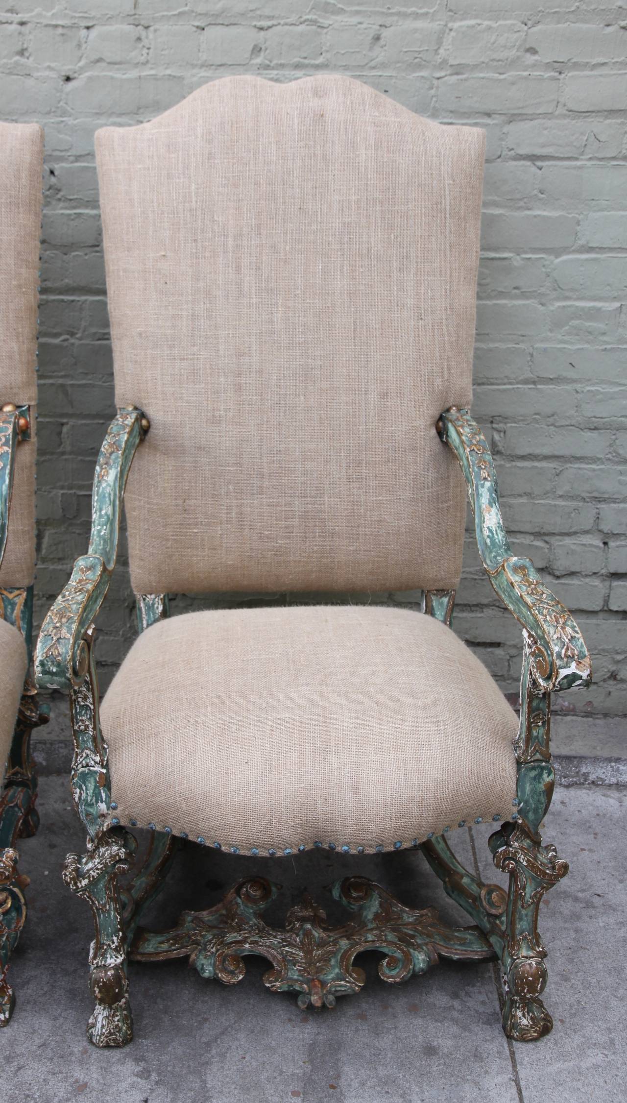 Pair of Italian Baroque painted and parcel-gilt armchairs each upholstered in burlap textile with nailhead trim detail and having a carved and green painted frame, high back, scrolling arms and an elaborately carved scrolling H-shaped stretcher.