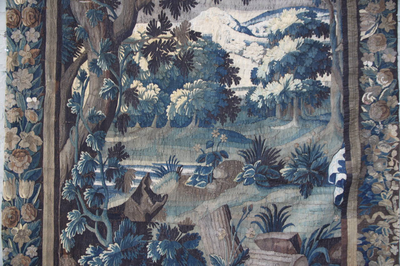 18th century handwoven Flemish tapestry of landscape done in beautiful shades of blue and brown depicting a winter woodland scene with snow on the mountains in the background and tree branches. Complete border depicting a wide range of flowers and