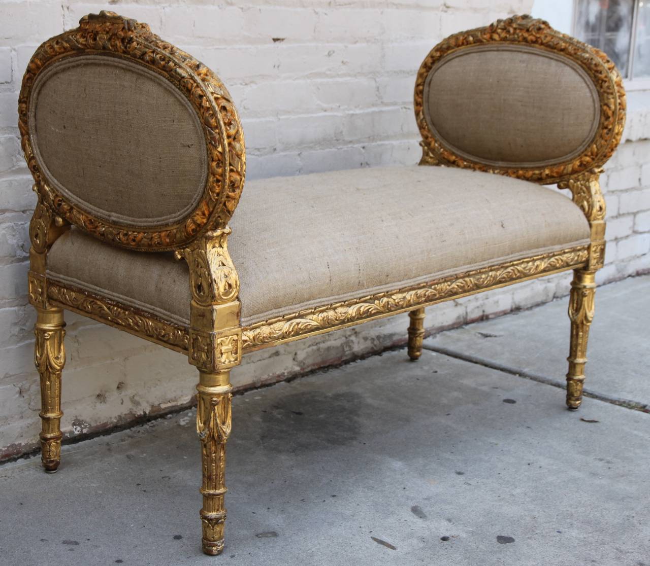 19th century heavily carved giltwood French neoclassical style music bench with oval-shaped sides. The bench is newly upholstered in burlap textile with self cording. The piece stands on four straight fluted legs with carved garlands. Carved detail