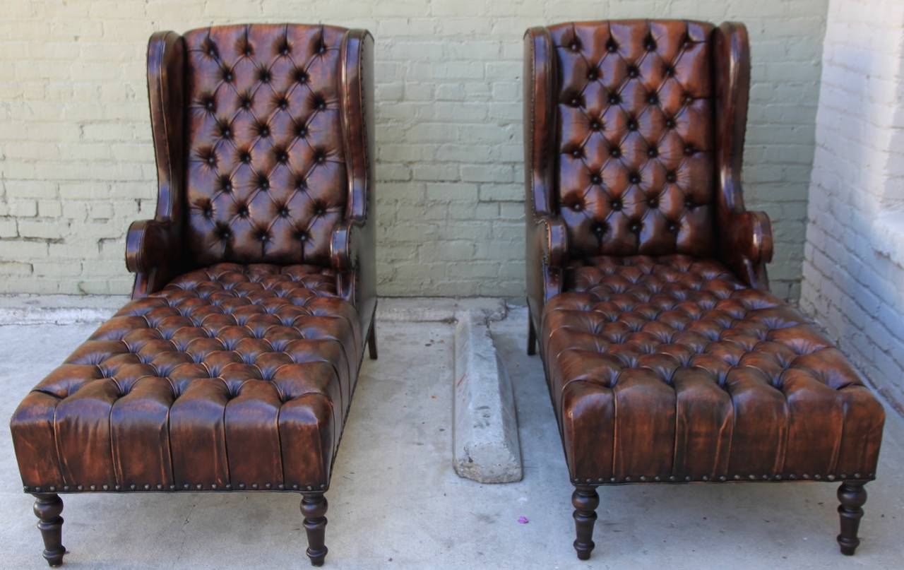 Pair of unusual handsome leather tufted wingback chaises with nailhead trim detail. The chaises stand on four legs stained in a similar coloration. Self cording and flat welt finished with large spaced nailhead trim detail.