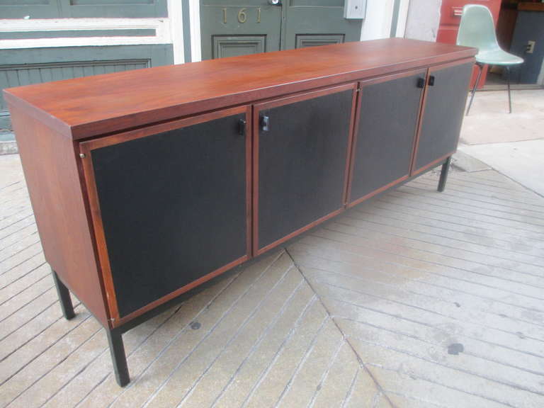 Large walnut credenza designed by John Kapel for John Stuart with four black vinyl covered doors which reveal one large adjustable shelf on the right and two pull out drawers and storage on the left.  Base is ebonized wood and .handles are painted