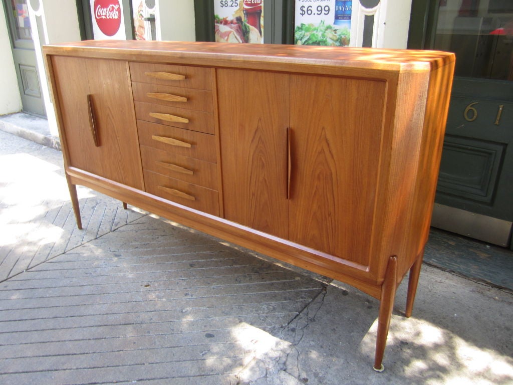 Large teak buffet of angled corners,6 center drawers and two sets of hinged doors supported by cradle of solid wood on four legs.  Branded UM marking. Johannes Anderson did many of the UM designs
