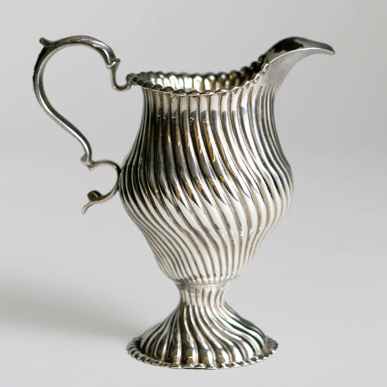 Finely detailed silver creamer in a ribbed swirl pattern with graceful curved handle and pedestal base. Indistinct hallmarks but with clear European hatchet mark.
