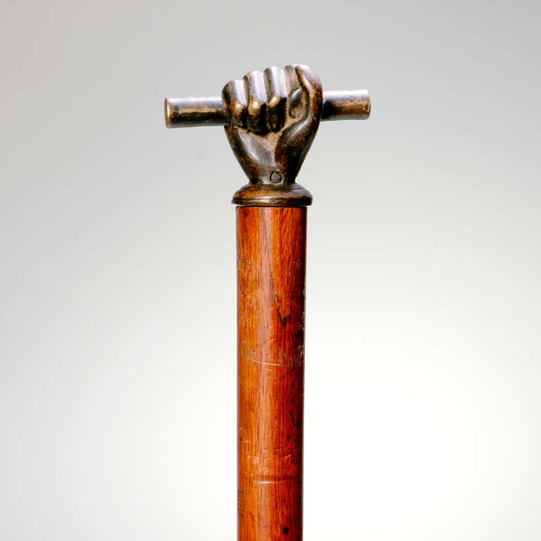 Wooden cane with bronze corkscrew handle in the shape of fist grasping a baton. Corkscrew unscrews from cane for use.