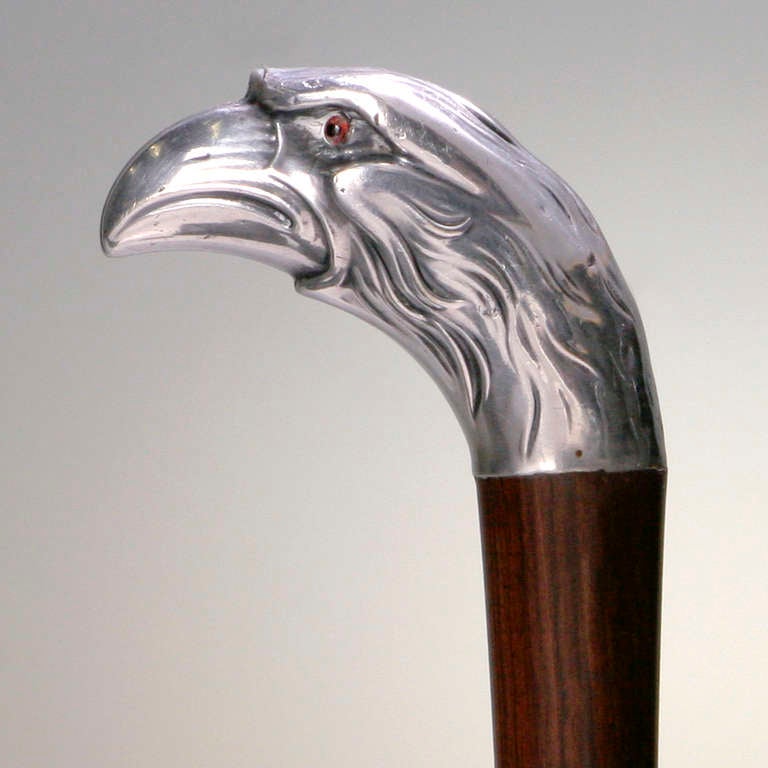 Wood and silver cane with striking eagle head as handle. Bird of prey has small red glass eyes and piercing stare. Stamped: Alpacoa.