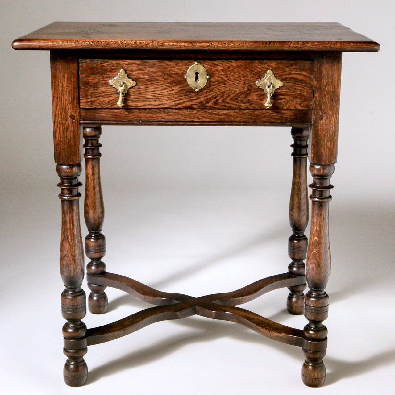 Traditional polished English oak side table with cross stretcher and turned legs. The table features a long drawer with lock and key. This Classic table is handcrafted in English workshops using the finest materials and antique tools to recreate a