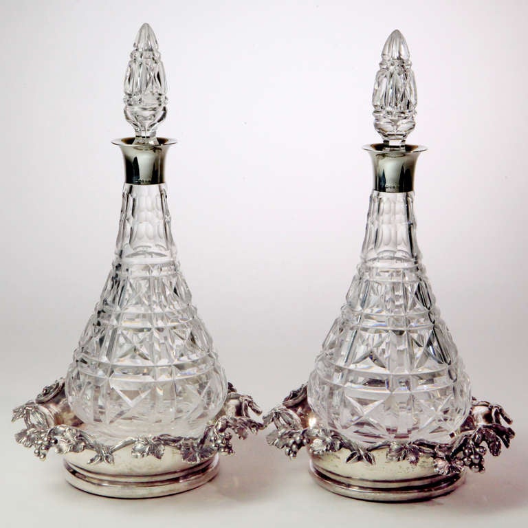 A notable pair of 19th century Edwardian cut crystal wine decanters with hallmarked silver mounts and tall crystal sphere stoppers. Together with complementary silver wine coasters with embossed grape cluster and leaf design.