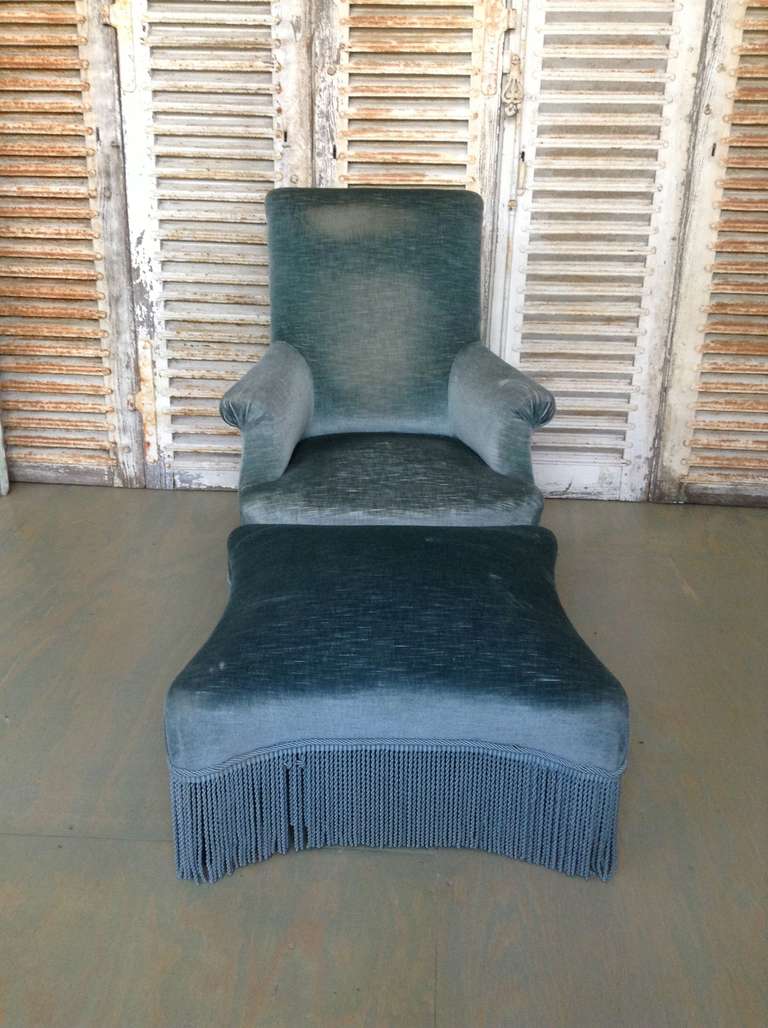  French 19th armchair and ottoman, scrolled back and rolled arms, upholstered in rich blue velvet and matching bouillon fringe. SOLD AS IS.