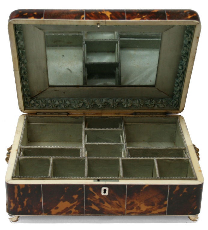 This beautiful tortoise jewelry box has a hinged top with inlayed tortoise panels. The top opens to display 12 lined compartments and a looking glass. On either side are lion ring handles and it sits on ball feet. A beautiful tortoise box to keep