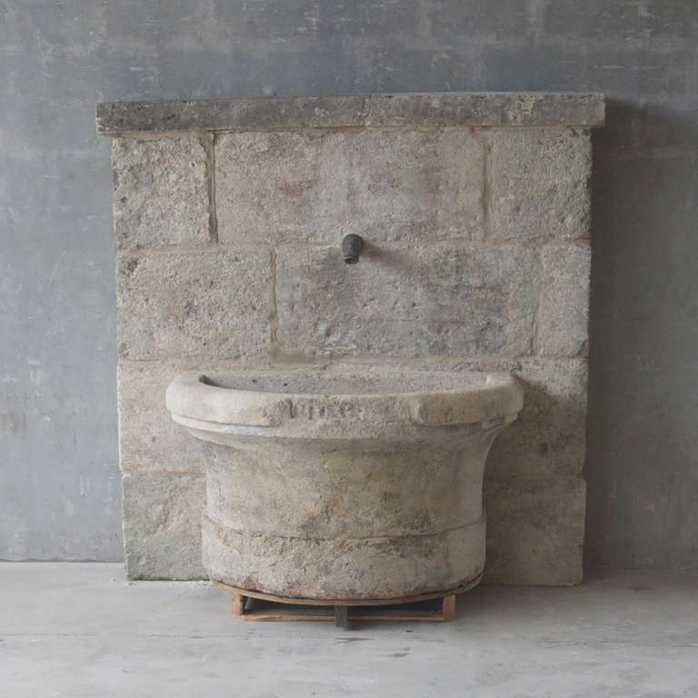 This 18th century Fountain was reclaimed from a Maison Bourgeoise in Grignan, France. Featuring a flat stone wall as a back and a rounded basin, this unique stone fountain is rustic. The antique animal-shaped fountain spout adds an element of charm.