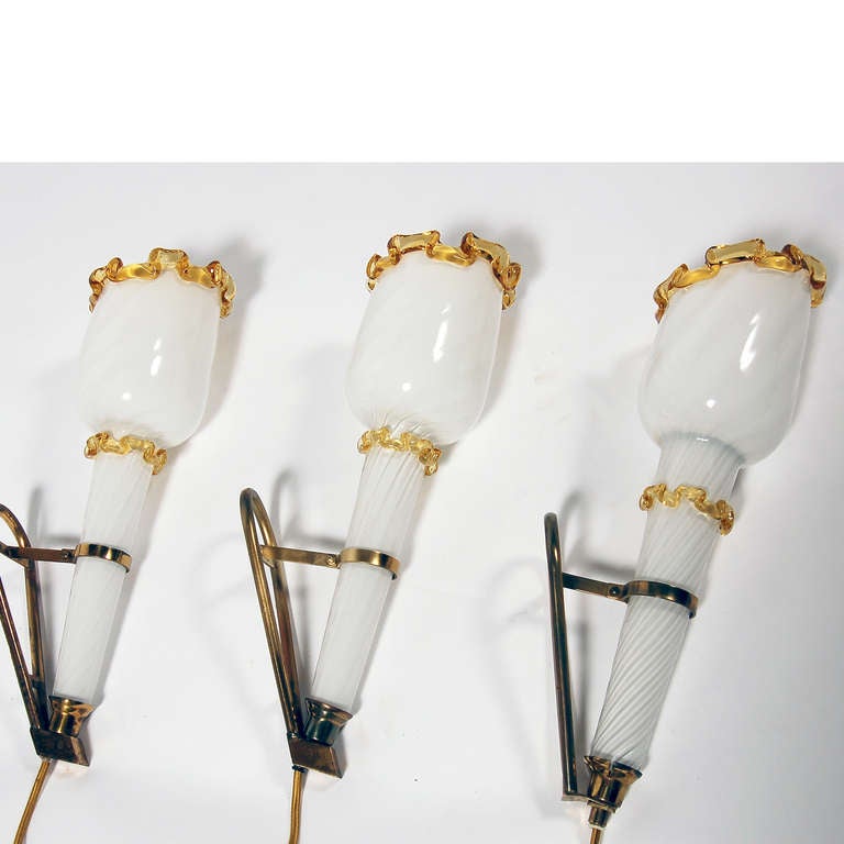 Three wall sconces in lattimo glass with amber decorative trim on brass wall attachments. Two matching, one with different connection at bottom and lower glass trim. Originally from movie house in Halsningland, Sweden.