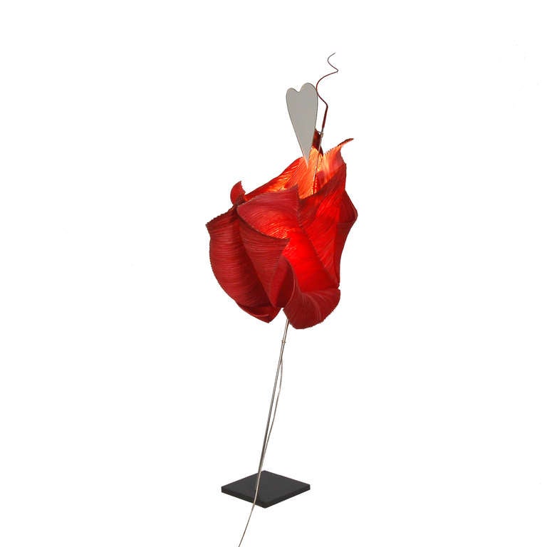 This table lamp is from the series The MaMo Nouchies by Ingo Maurer. The shade is red paper, which is curved around a bar of stainless steel. The end of the lamp bar is configured by a red silicone peak, underneath a heart shaped mirror is located