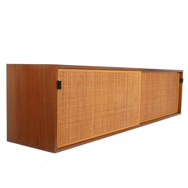 Wall mounted credenza with walnut case and cane front sliding doors with leather tab pulls.  Adjustable interior shelves made of oak. Retains early label.