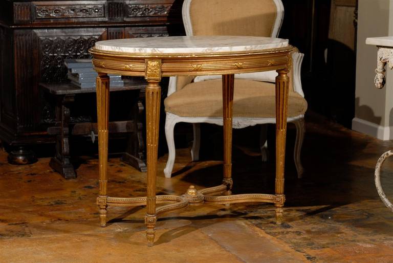A French Louis XVI style giltwood oval centre table with white variegated marble top, fluted tapering legs and curved X-form cross stretcher from the 19th century. This French centre table features a white variegated marble top of oval shape,