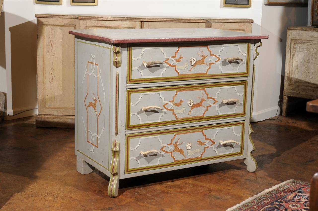 An English vintage three-drawer commode with painted swags and antlers hardware from the turn of the century. This painted commode features a slightly sinuous rectangular top adorned with a geometrical cartouche receiving in its center two leaping