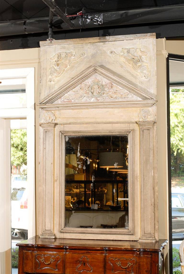 This exquisite French painted wood trumeau mirror from the 19th century features an elegant classical frame, reminiscent of a Greek or Roman temple. The eye is immediately drawn to the triangular pediment, flanked with two graceful angels, with one