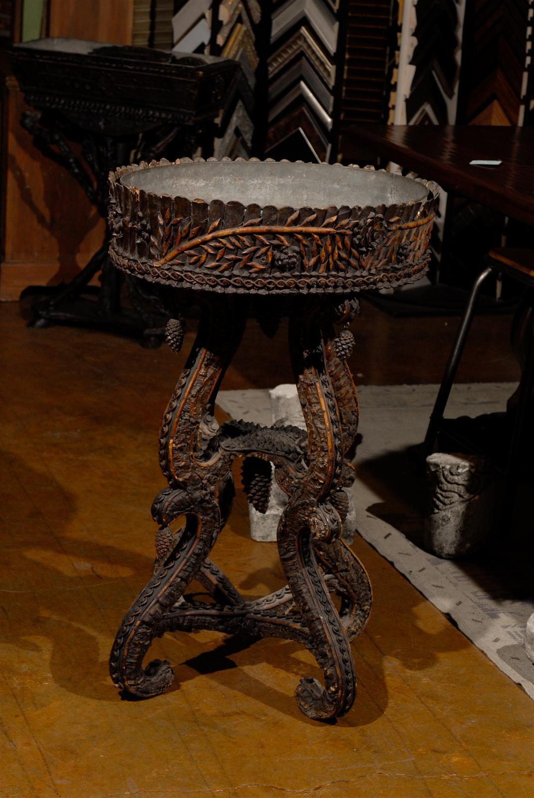 19th century jardiniere, the circular top with an inset metal conforming basin, raised on four scrolling legs joined by a curved X-form stretcher, with all over forest themed carving, including hanging pine cones throughout. 