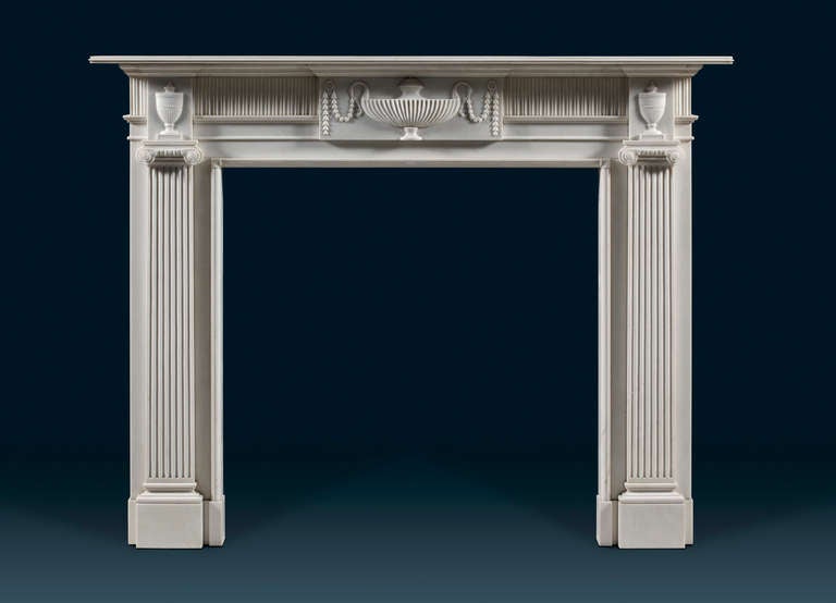 This Irish chimneypiece of the Greek ionic order features a central tablet carved with a ribbed classical tazza shaped vase draped with swags of flowers, housed in a fluted frieze. The corner blocks are adorned with lidded urns and sit above jambs