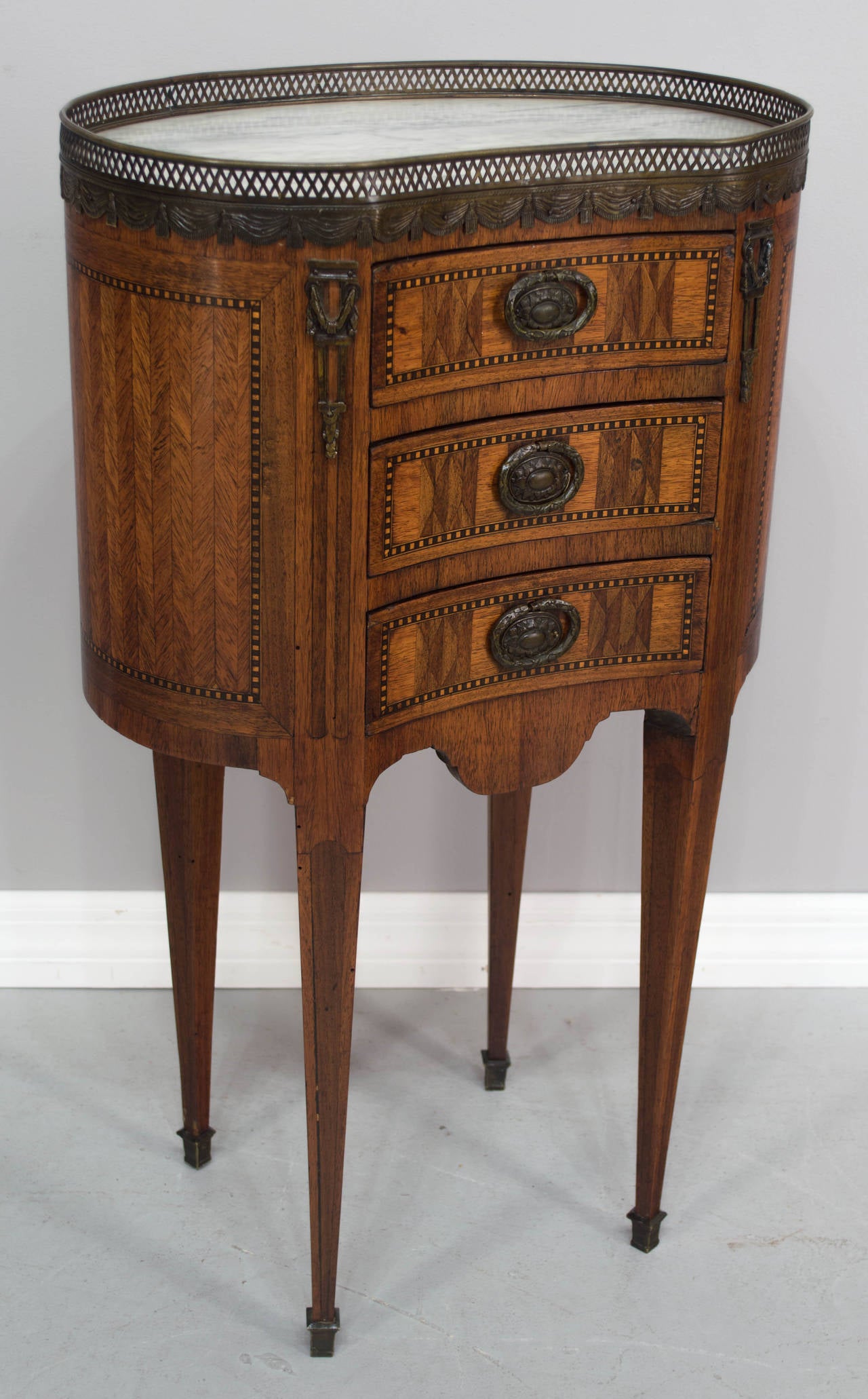 19th century French Louis XVI style marquetry side table in an unusual kidney shape with slender tapered legs. Made of veneer of mahogany and walnut with three dovetailed drawers, marble top surrounded by bronze gallery. Original patinated bronze