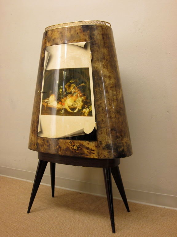 This is a rare and unusual bar cabinet by Aldo Tura and is probably one of a kind. It is a triangular oval shape, made of wood and covered in goat skin or parchment with a Fornasetti style trompe l'oeil design on the door depicting a vibrantly