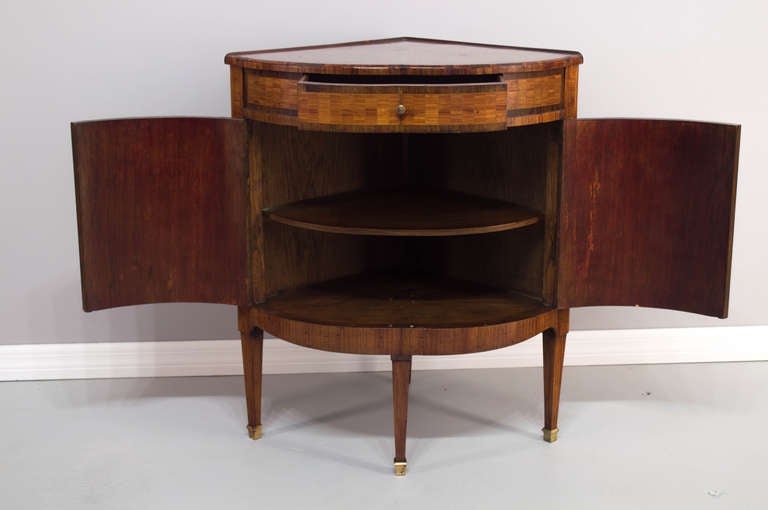 A small corner cabinet with two curved doors below a center drawer, inlay of mahogany and walnut and cherry, opening with one inside shelf, on four tapered legs. Note that the piece slant a bit to the back for stability. Bronze sabots and brass