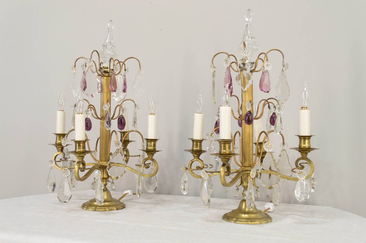 An elegant and whimsical pair of French Girandoles in brass.  These lamps, composed of turned and fluted brass are adorned with faceted clear and violet crystals.  Finishing the design is a geometric glass finial. In working order. Rewired.