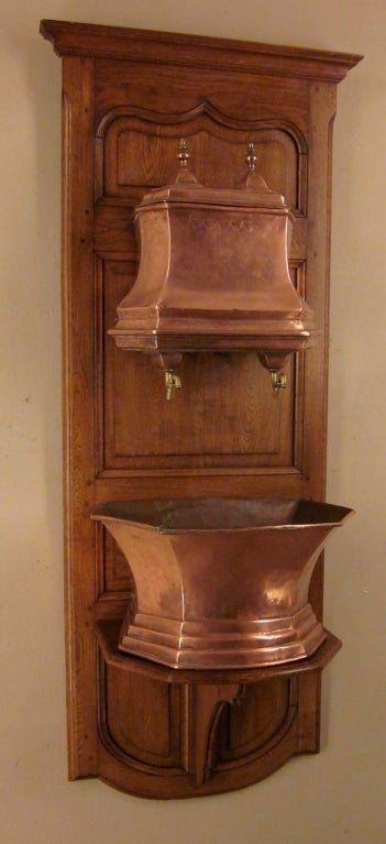 An 18th century copper fountain with lavabo and brass faucets mounted on a later oak panel with raised panels and pegged construction.