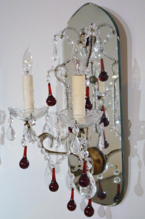 A pair of two-light French sconces mounted on mirrored backplates with beveled edges. The arms are outlined with crystal beads and draped with clear and red teardrop crystals.