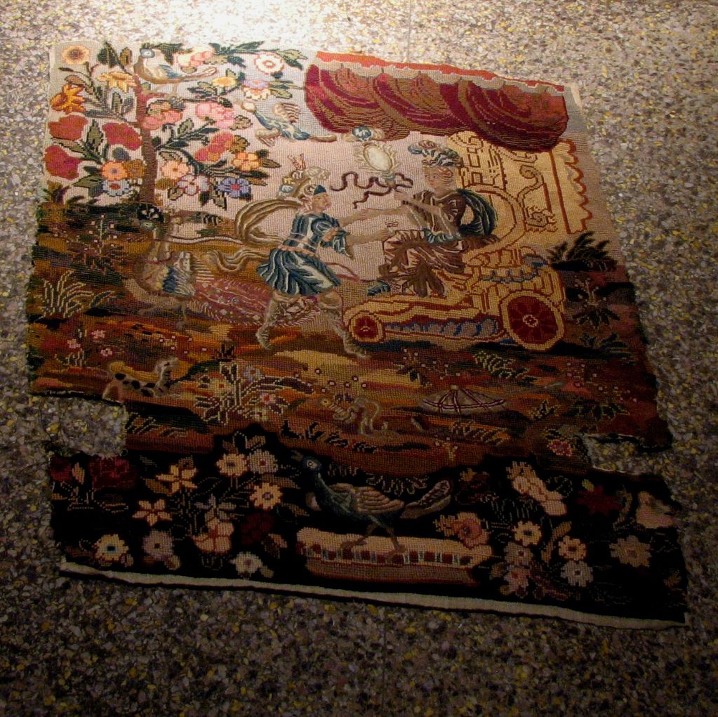 AN ANTIQUE ENGLISH MID-EIGHTEENTH CENTURY, NEEDLEWORK PANEL, DEPICTING A ROYAL PERSON HOLDING A SCEPTER, BEING DRAWN IN A COVERED CHARIOT BY A MYTHICAL BEAST WITH A SOLDIER IN ATTENDANCE, WITH DOG, BIRD IN FLIGHT,AND PEACOCK