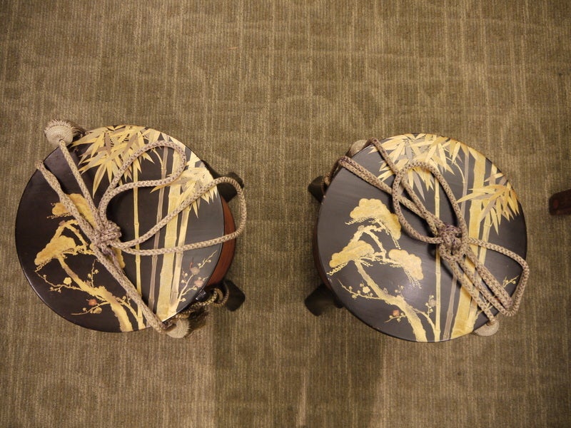 A pair of round Japanese lacquer storage boxes with lids.  Legs are black lacquer.  Tops have black background, and gold bamboo design.