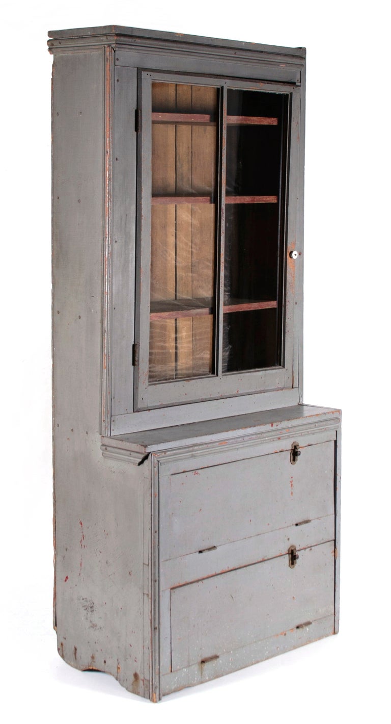 NARROW, MAKE-DO PENNSYLVANIA CUPBOARD IN OLD GREY PAINT OVER AN EARLIER RED, WITH A GLAZED DOOR AND TWO UNUSUAL DROP-DOWN DOORS, CA 1890:

Unusual country step-back cupboard with plank sides on shallow, half-moon, boot-jack feet; a make-do form,