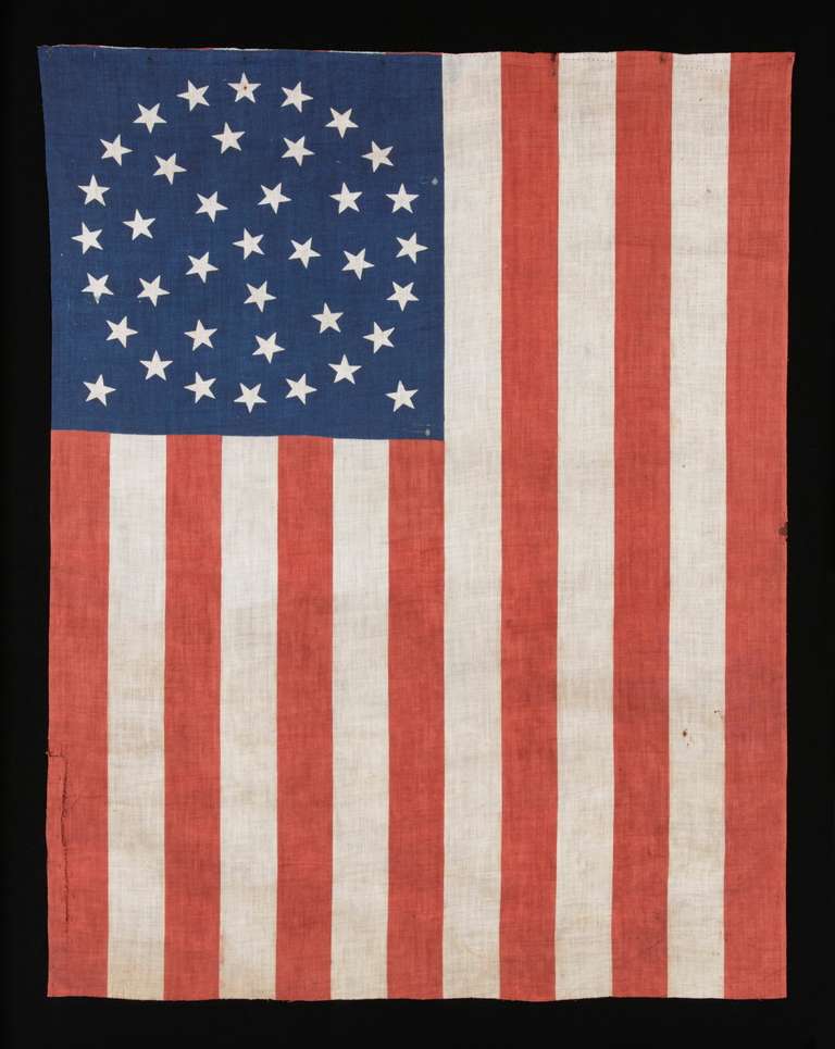 38 star American parade flag, printed on plain weave cotton. The stars are arranged in a triple-wreath form of the medallion configuration, with a single center star and two stars flanking outside the basic pattern toward the fly end. Typically