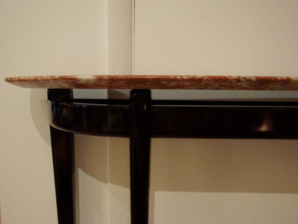 A mid century demi-lune console table featuring a frame in ebonised Mahogany with four shaped tapering legs, a one piece four armed stretcher and a lowered bent wood skirt. The base supports a top of figured marble in shades of red, orange and amber