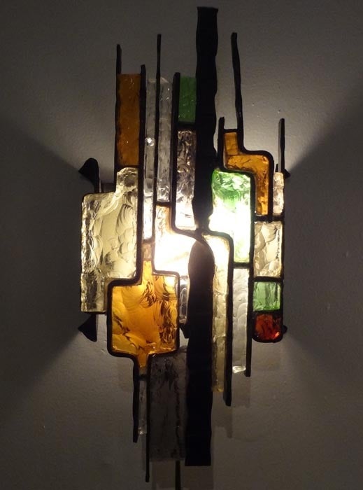 A pair of sculptural wall sconces in the brutalist style each featuring a frame in darkened steel designed in an abstract modernist pattern with insets of hand chipped glass which act as a light diffusing shade for the interior light source. In the