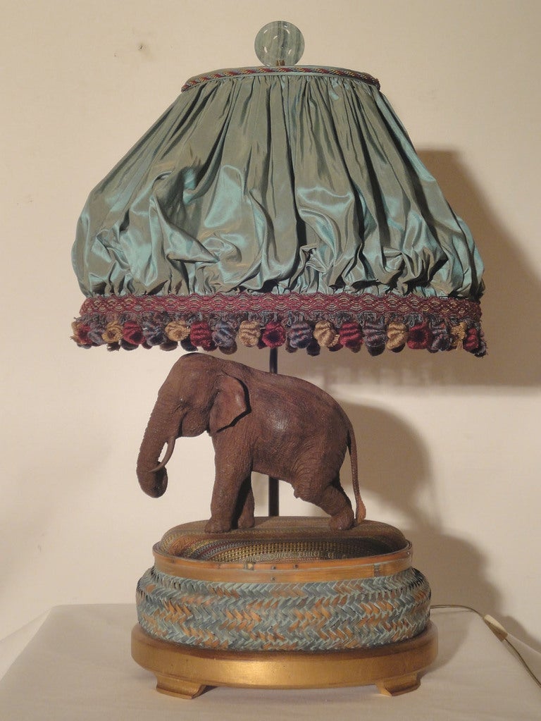 Pair of Well Carved Wooden Elephants From India are mounted on a fabric covered base featuring a woven stripe. An oval basket weave base with blue accent paint rests on a simple gilt stand with feet. The custom balloon-petticoat silk shade with