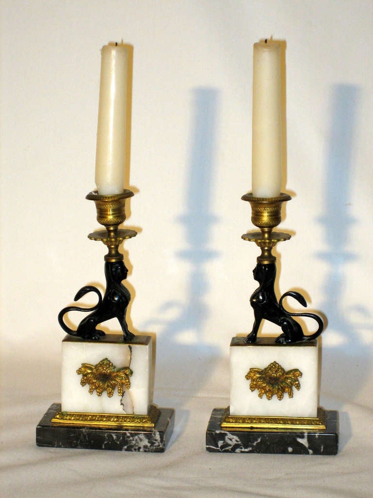 A petite pair of Empire Style Candlesticks in the form of a Black Lion Sphinx sitting upon a white onyx plinth mounted with gilt bronze.  The base is a white and charcoal gray marble.  The candle nozzles and bobeches are detailed with a classical