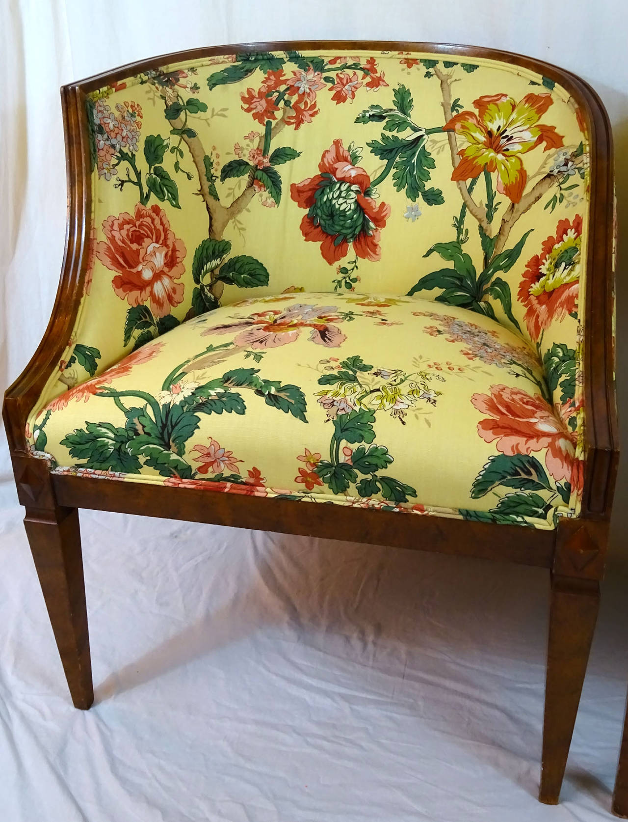 Left and right pair of stained beechwood portrait chairs, one side has a high pointed arm and one side has a lower curved arm. Upholstered in a yellow floral fabric with pink, blue, and yellow flowers, amongst green vines and leaves.