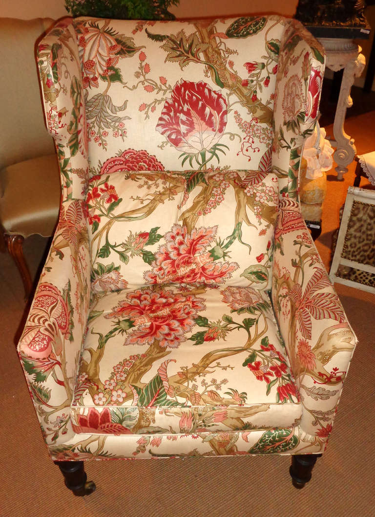 19th century English Wingback chair upholstered in cream, red, and green with removable back pillow, sitting upon mahogany legs with brass casters.