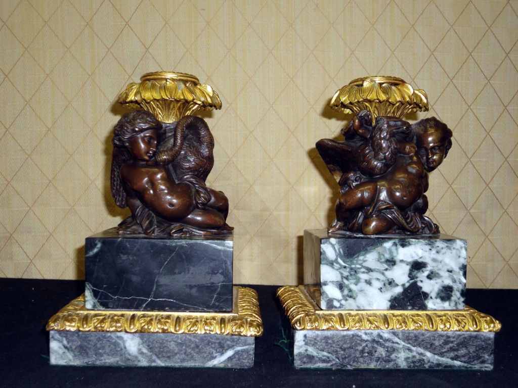 Pair of Bronze & Doré Putti Candlesticks with Eagle & Swan on Rich Dark Green Marble Bases. The small patinated sculptures depict figures of mythological significance. The plinth base is surrounded by a lovely gilt bronze molding & the candle nozzle