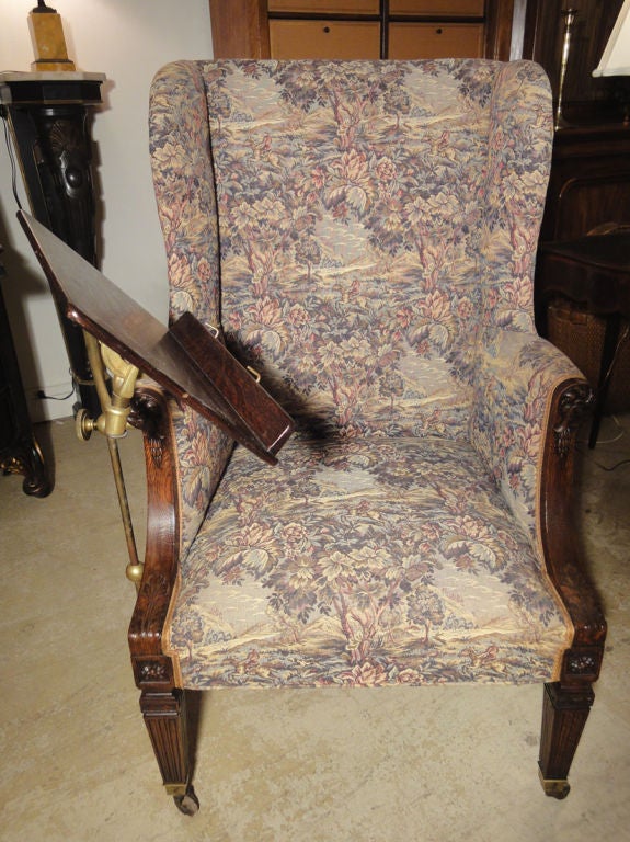 Sheraton Style Wing Chair on Casters with Attached Reading Stand. The tall upholstered wingback rests above slightly rolled arms that feature carved exposed uprights of decorative pateras & acanthus leaves. The tapered stop-fluted legs end in brass